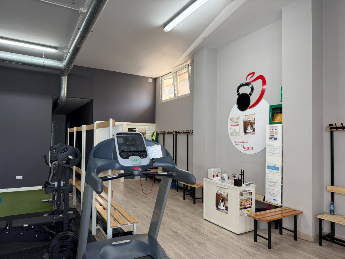 Interior de gimnasio con cinta de correr, bancos de madera y logo de pesa rusa en la pared para entrenamiento personalizado - Saludx10